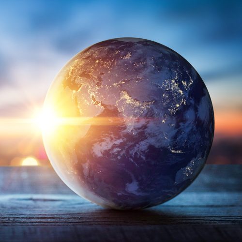 A globe of Earth resting on a textured wooden surface, with a blurred cityscape and a bright sunset behind it.